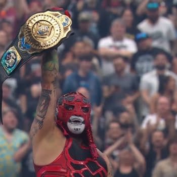 A masked wrestler in a vibrant red outfit triumphantly holds up a championship belt above their head in front of a cheering crowd. The background shows enthusiastic fans celebrating the moment.