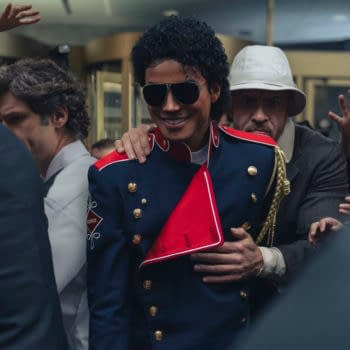Jaafar Jackson portrays Michael Jackson in a blue military-style jacket with a red accent, surrounded by a crowd. The scene captures the intensity of a moment with hands reaching out, conveying a sense of excitement.