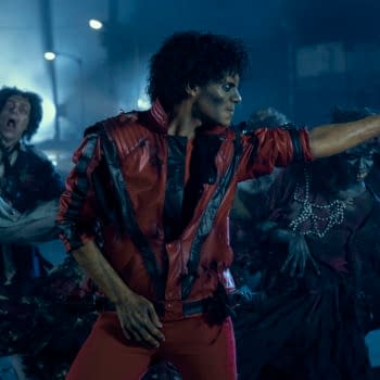 Jaafar Jackson portrays Michael Jackson in a dynamic scene, wearing a distinctive red jacket with black details, surrounded by a group of zombie-like figures. The atmosphere is moody and dark, reflecting a dramatic moment from the upcoming biopic 'Michael'.