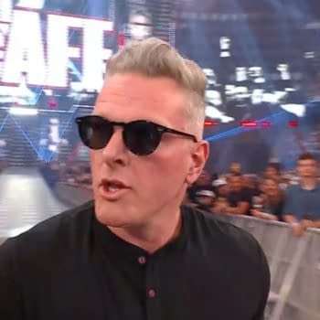 A man with short, silver hair wearing sunglasses walks past a crowd at a wrestling event, with bright lights and a large screen in the background displaying 'The McAfee'.