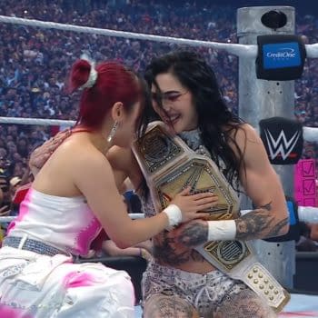 Two women embrace joyfully in a wrestling ring, one holding a championship belt. The crowd is visible in the background, celebrating the moment.