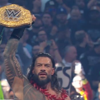 A victorious wrestler holds the WWE championship belt aloft, adorned in traditional attire with long hair and a serious expression, set against an enthusiastic crowd at a wrestling event.