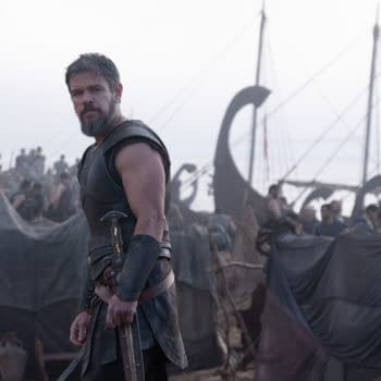 Matt Damon as Odysseus stands in armor at a crowded ancient Greek encampment beside a ship.