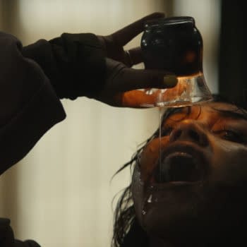 A tense horror scene from Evil Dead Burn shows a woman tipping a dark jar and pouring liquid onto her face in a dimly lit room.