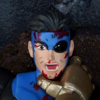 Close-up of a battle-damaged Invincible action figure with a blue-and-black costume, blood marks, and one damaged eye. The figure appears posed in a gritty, dramatic scene.