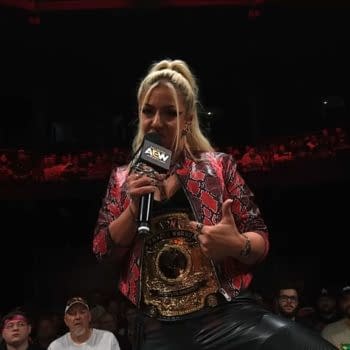 Thekla speaks into an AEW microphone while wearing a championship belt and patterned jacket in a dimly lit arena. She appears to be cutting a promo before Forbidden Door on AEW Collision Summer Blockbuster.