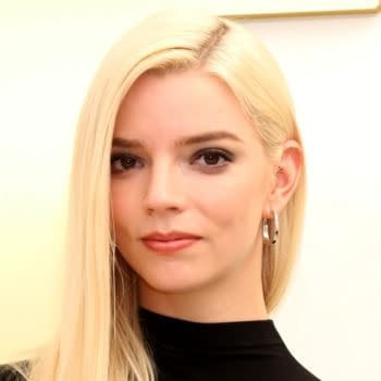 Anya Taylor-Joy poses at an Apple TV press event, wearing a black outfit and silver hoop earrings. Her platinum blonde hair is parted to one side against a bright backdrop.