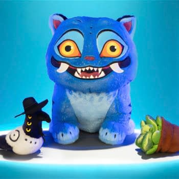 A blue Derpy Tiger plush from KPop Demon Hunters sits on a glowing display with two small companion figures. The plush has oversized eyes, sharp teeth, and a playful, cartoonish look.