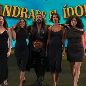 Andrade El Idolo walks to the ring at AEW All In: London with four women in black dresses beside him. A bright tropical-style entrance screen fills the background.