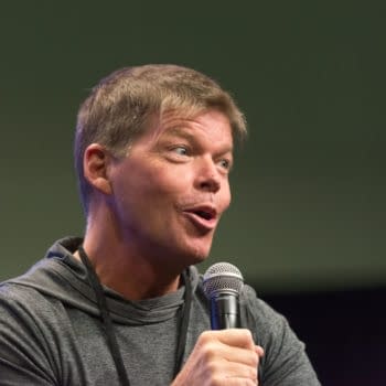 Rob Liefeld speaks into a handheld microphone at a comic convention panel in Los Angeles. He wears a gray hoodie and looks animated mid-sentence.
