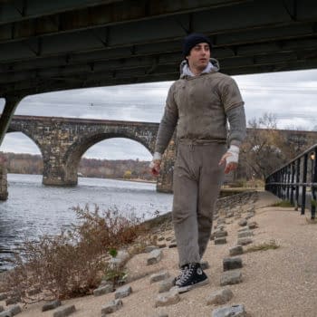 Anthony Ippolito, as Sylvester Stallone in I Play Rocky, walks along a riverside path beneath a bridge, wrapped in a gray hoodie and sweatpants with bandaged hands.