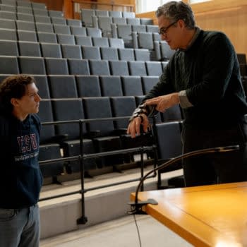 Two men confer in a lecture hall, one showing a wrist device or prosthetic arm to the other. Rows of empty auditorium seats rise behind them.