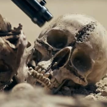 A grim close-up of a buried skull in the sand, with a dark firearm barrel aimed at it, from the Crossed movie teaser.