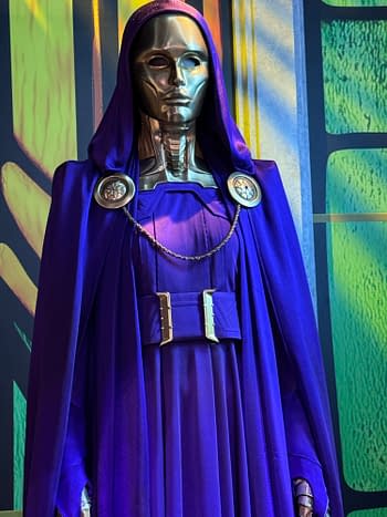 A purple-hooded metallic masked figure in an Avengers: Doomsday costume display at SDCC, standing against a colorful comic-style backdrop.