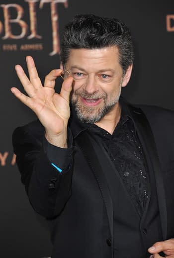 Andy Serkis, dressed in a formal black suit with a patterned shirt, smiles while holding up a small golden ring, presumably referencing his role in "The Hobbit," at a movie premiere event.