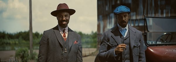 Two men stand outdoors, both dressed in stylish vintage suits. One man wears a maroon fedora, while the other sports a blue cap, with a classic automobile visible in the background.