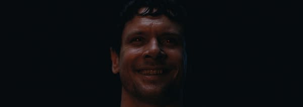 A close-up portrait of a man with a dark background, featuring a slight smile and intense expression, illuminated by soft light that highlights his facial features.