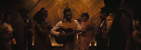 A lively scene depicting a group of diverse individuals gathered in a dimly lit setting, with a man playing guitar in the foreground. The atmosphere conveys a sense of celebration and community, as people engage and enjoy the music.
