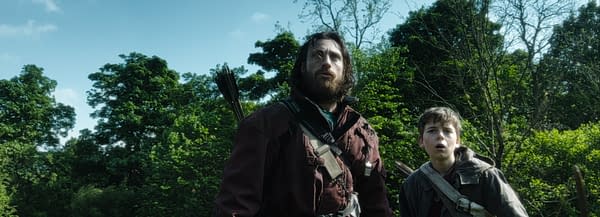 A dramatic scene featuring a bearded man and a young boy in a forested area, both showing expressions of surprise or fear. The man is wearing a dark jacket with arrows on his back, while the boy is in a simpler outfit, indicating a tense moment in a narrative.
