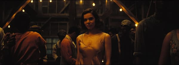 A scene in a dimly lit gathering, featuring a woman in a light dress standing prominently amidst a crowd. Warm ambient lights hang above, casting a cozy atmosphere as the people around her engage with each other.
