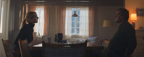 A tense scene from the film 'Over Your Dead Body' featuring two characters seated at a wooden table in a warmly lit room, facing each other with intense expressions.