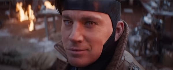 Channing Tatum is depicted in a cinematic scene, wearing a dark headpiece and a brown jacket, with a confident expression as flames flicker in the background.