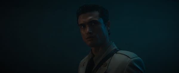 Charles Melton stands in a dimly lit, moody scene wearing a light-colored uniform or suit, with his face partly shadowed and turned toward the camera.