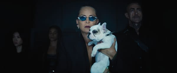 A woman in a black suit holds a small white French bulldog, wearing sunglasses. She is flanked by two figures in dark clothing, against a dimly lit background.