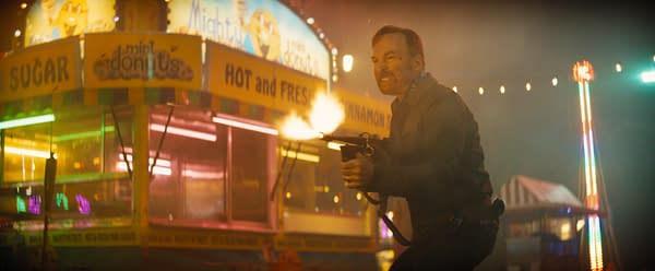A man in a dark jacket holds a firearm and looks determined amidst a carnival setting, with bright lights and food stands featuring signs for mini donuts and sugar. The scene conveys intensity, possibly during an action sequence.