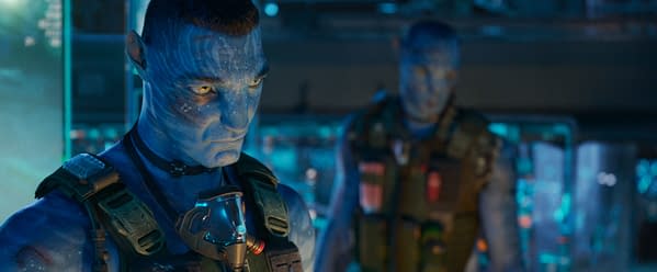 Quaritch, portrayed by Stephen Lang, appears in a tense moment in the film 'AVATAR: FIRE AND ASH,' showcasing his blue skin and tactical gear against a futuristic backdrop filled with ambient lighting.