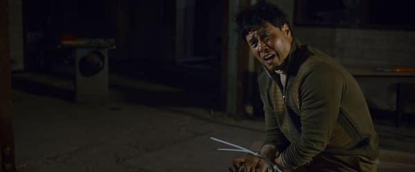 A distressed character, portrayed by Randall Park, sits on the floor in a dimly lit setting, his hands bound with plastic ties, conveying a sense of tension from the film 'Eenie Meanie' by 20th Century Studios.