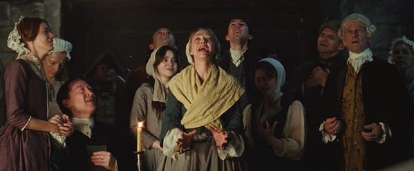 A scene from 'The Testament of Ann Lee' featuring multiple characters in historical attire, some expressing strong emotions as they gather around a candle. The atmosphere suggests a dramatic moment, possibly involving themes of faith or revelation.