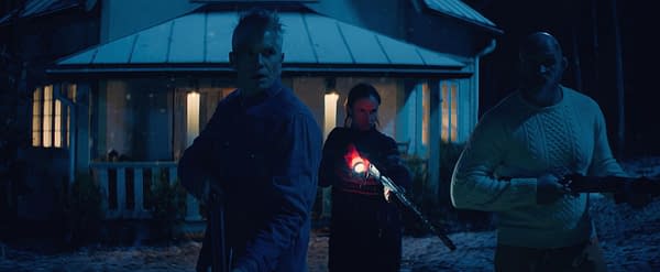 A dramatic scene from the film 'Over Your Dead Body', featuring three people standing outside a house at night, each holding firearms. The atmosphere is tense, illuminated by the house's warm lights contrasting with the dark surroundings.