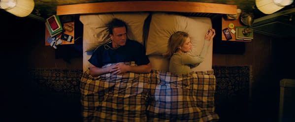 A still from 'Over Your Dead Body' depicts two people in an intimate setting, lying in bed with a plaid blanket. They appear to be awake but distant from each other, surrounded by bedside items.