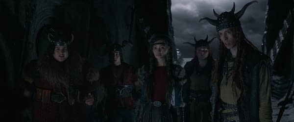 A group of young Viking characters stands together in a dimly lit, rocky environment, each adorned with distinct outfits featuring horns and fur elements. Their expressions convey a sense of curiosity or concern, fitting the adventurous theme of the 'How To Train Your Dragon' series.
