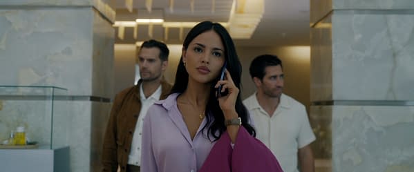A woman in a light purple blouse is speaking on her phone, with a serious expression. She is accompanied by two men in the background, one wearing a brown jacket and the other in a beige shirt, in a modern, elegant setting.