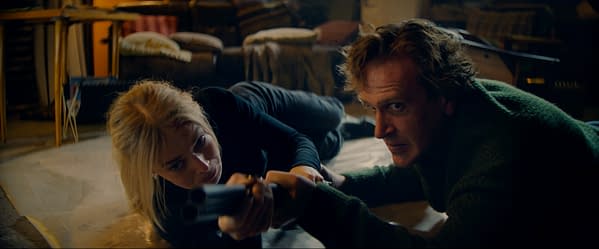In a tense scene from the film 'Over Your Dead Body', a woman and a man are lying on the floor, both appearing focused and determined, with the woman aiming a gun. The background suggests a cluttered room with furniture and soft furnishings.
