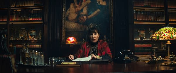 Anjelica Huston portrays The Director in a scene from 'Ballerina', seated at a desk in a richly decorated room filled with books and antiques. A dimly lit stained glass lamp and various objects adorn the desk, creating a dramatic atmosphere.
