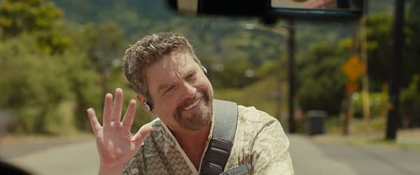 Zach Galifianakis as Jumba in Disney's live-action 'Lilo & Stitch', smiling and waving, wearing a patterned shirt. The background features a street scene with greenery and road signs.