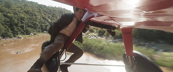A scene from 'Mission: Impossible - The Final Reckoning,' featuring a tense moment where a man hangs onto a small red aircraft, with a lush green landscape and a river visible below.