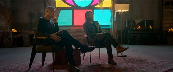 Chad Stahelski and Keanu Reeves sit in a dimly lit room, surrounded by candles and a vintage piano, with colorful stained glass in the background. The atmosphere suggests a contemplative moment, possibly reflecting the themes of 'Wick is Pain'.