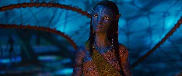 A still from 20th Century Studios' 'Avatar: Fire and Ash', featuring Neytiri, a blue-skinned character with long braids, wearing a woven top and a patterned sash. The scene is set against an otherworldly, illuminated blue background.