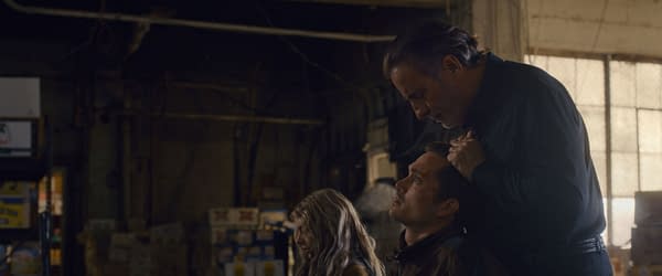 A tense scene from the film 'Eenie Meanie' featuring Samara Weaving as Edie, Karl Glusman as John, and Andy Garcia as Nico in a dimly lit industrial setting. The characters display intense emotions, with Nico holding John's head, suggesting a confrontation.