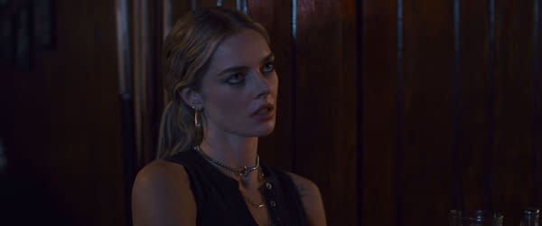 Still from the film 'Eenie Meanie' featuring Samara Weaving as the character Edie, shown seated in a dimly lit bar with wooden paneling in the background.