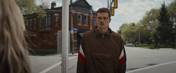 A scene from 'Eenie Meanie' featuring Karl Glusman as the character John, standing on a street corner, wearing a brown jacket with red and white stripes. A vintage brick building and traffic signals are visible in the background.