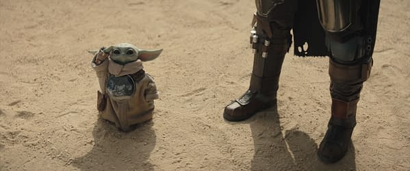 Grogu stands in the foreground, wearing a simple cloak and making a playful gesture, while The Mandalorian stands beside him, dressed in his iconic armor and cape. The sandy background highlights the duo's adventurous setting from the series.