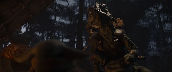 Grogu faces a creature in a dark, forested setting from Lucasfilm's 'The Mandalorian and Grogu.' The creature appears to be wearing a vest and has a textured, reptilian appearance.