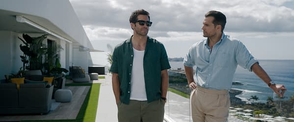 Two men are standing outdoors on a modern balcony overlooking the ocean, dressed in casual, stylish outfits. The scene conveys a relaxed atmosphere with a contemporary setting and lush greenery.