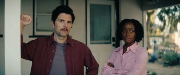 A still from the film "The Saviors," featuring a man with a mustache wearing a maroon shirt, leaning against a post, and a woman with curly hair in a pink button-up shirt, standing with her arms crossed, both appearing serious against a blurred outdoor background.