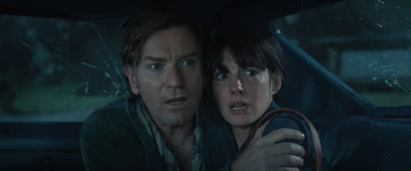 A tense movie still shows a man and woman huddled together in the back seat of a car, staring fearfully ahead through a shattered window.
